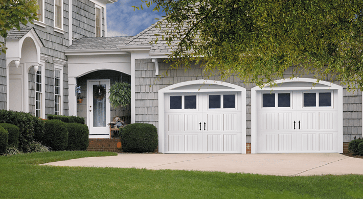 Suburban house with gray shingle siding and two white garage doors with four windows on the top panel of each door